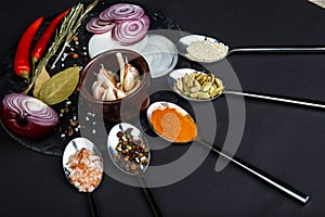 A board with onion, garlic, bay leaf, pepper and salt beside which lie spoons with various spices.