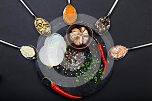 A board with onion, garlic and pepper around which spoons with spices. Top view on a black background.