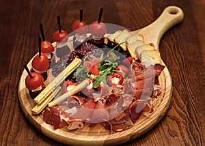 Board with snacks on wooden table. Cold appetizers with tomatoes, sausage, salami, ham, arugula, olives and bread