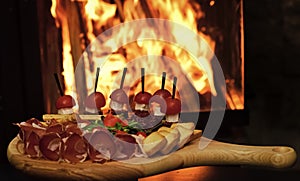 Board with snacks on wooden table, fire on background. Cold appetizers with tomatoes, sausage, salami, ham, arugula