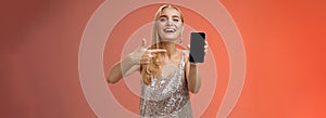Boastful proud charming elegant blond woman in stylish evening dress show smartphone display proudly pointing mobile