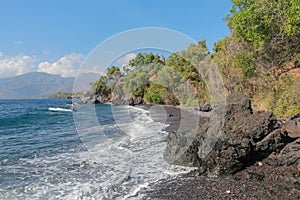 Boat anchored at the beach with black sand and lava rocks. Dense oil vegetation and mountain range on background with white clouds