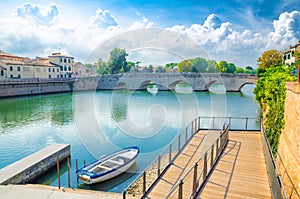 Boat in blue water near pier and stone arch Tiberius bridge Ponte di Tiberio, Augustus Bridge over Marecchia river in Rimini