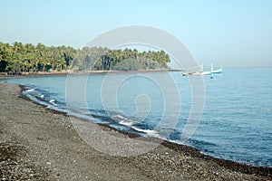 A boat on the sea with pebbly shore surrounded by tropical trees in an island under a blue sky