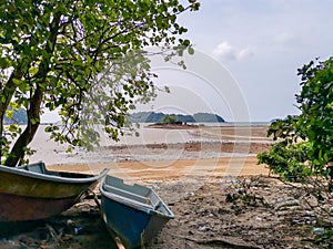 Boat and tree foreground and small tropical island in the middle of sea and cloudy blue sky background at low tide period