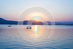 Boats floating on Lake Pichola with colorful sunset reflated on water beyong the hills. Udaipur, Rajasthan, India.