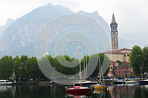 Boats on lake Como near church with tower, trees and mountain in background.