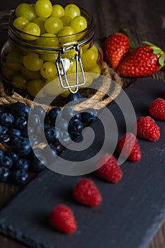 Bodegon fruit, fresh strawberries on slate plate, blueberries and glass jar with fresh grapes on an aged basis