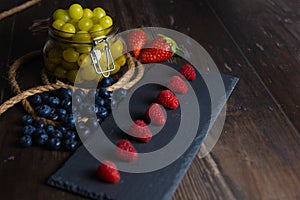 Bodegon fruit, fresh strawberries on slate plate, blueberries and glass jar with fresh grapes on an aged basis photo