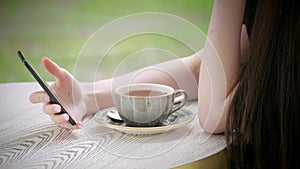 Body part hands of young Caucasian girl holding smartphone in hand. Cup of tea on table coffee terrace