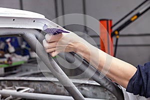 The body repairman grinds the white car`s frame with purple emery paper in preparation for painting after applying putty in a photo