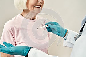 Body temperature. Doctor in blue sterile gloves using electronic thermometer for measuring temperature of senior female