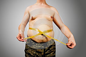 Body of young man with excess weight, keep belly fat, measure itself with a measuring tape on gray background