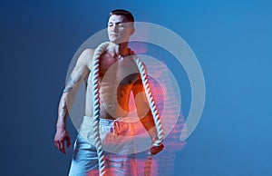 Bodybuilder with bared torso posing with the rope in studio on blue background.