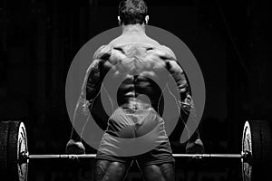 Bodybuilder with great physique and body shape training with barbell in gym over dramatic light