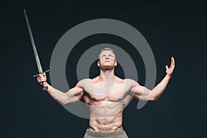 Bodybuilder man posing with a sword isolated on black background. Serious shirtless man demonstrating his mascular body