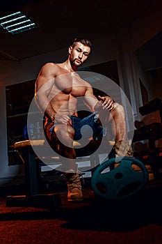 Bodybuilder sits on a weight bench, he takes a break. Muscular man at a workout place in a gym and smiling to camera.