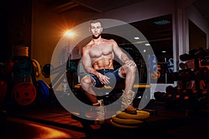 Bodybuilder sits on a weight bench, he takes a break. Muscular man at a workout place in a gym and smiling to camera.