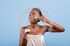 Bodycare concept. Pretty african american lady applying cream on her face, standing over blue background