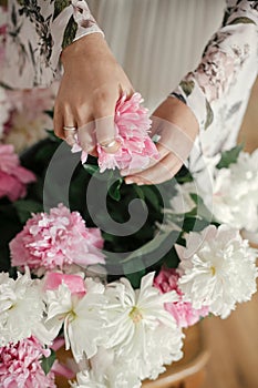 Boho girl holding pink and white peonies in hands at rustic wooden chair. Stylish hipster woman in bohemian dress arranging peony