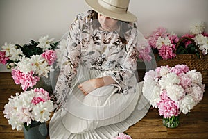 Boho girl sitting at pink and white peonies in rustic basket and metal bucket on wooden floor. Stylish hipster woman in bohemian