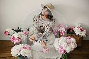 Boho girl sitting at pink and white peonies in rustic basket and metal bucket on wooden floor. Stylish hipster woman in bohemian