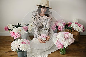 Boho girl sitting at pink and white peonies in rustic basket and metal bucket on wooden floor. Stylish hipster woman in bohemian