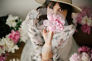 Boho girl sitting at pink and white peonies in rustic basket and metal bucket on wooden floor. Stylish hipster woman in bohemian