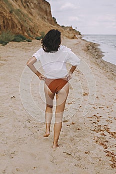 Boho girl in white shirt walking on sunny beach, back view. Carefree stylish woman in swimsuit and shirt relaxing on seashore.