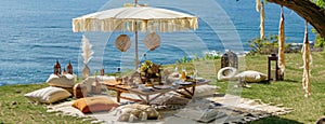 a boho picnic setup on lush grass near the ocean, featuring a cozy table set for two and a charming umbrella casting