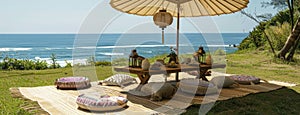 a boho picnic setup on lush grass near the ocean, featuring a cozy table set for two and a charming umbrella casting