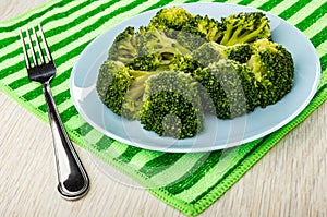 Boiled broccoli in blue plate, fork on striped napkin on wooden plate