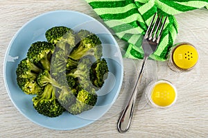 Boiled broccoli in plate, fork on napkin, salt and shaker on wooden plate. Top view