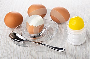 Boiled eggs, peeled egg, spoon on egg stand, salt shaker on wooden table