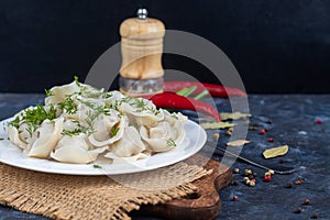 Boiled dumplings on a white plate. In the background is a wooden hodgepodge, greens, red peppers and bay leaves