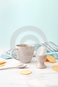 Boiled egg in ceramic egg cup, cup of coffee and thin crispy corn chips on background of turquoise kitchen towel. Breakfast