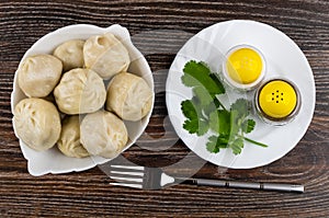 Boiled khinkali in bowl, plate with cilantro, salt, pepper, fork on dark wooden table. Top view