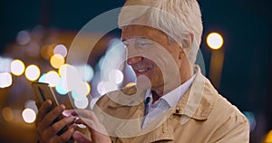 Bokeh shot of senior smiling man using smartphone on city street at night