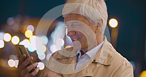 Bokeh shot of senior smiling man using smartphone on city street at night