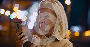 Bokeh shot of smiling aged lady standing on street at night replying to text on cellphone
