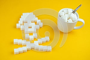 The bones skull made of sugar cubes and a white cup full of sugar with a spoon on a yellow background. Sugar kills and