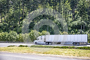 Bonnet white big rig semi truck tractor carry long dry van semi trailer moving on the highway road with green forest on the side