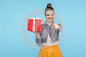 Bonus for you, winner! Portrait of optimistic beautiful fashionably dressed girl with hair bun holding gift box and pointing to photo
