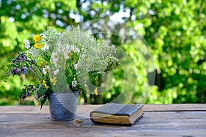 Book in black cover, bouquet of wild flowers, family bible lie on wooden table in garden, blurred natural landscape in background