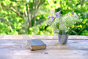 Book in black cover, bouquet of wild flowers, family bible lie on wooden table in garden, blurred natural landscape in background