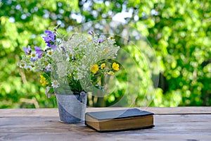 Book in black cover, bouquet of wild flowers, family bible lie on wooden table in garden, blurred natural landscape in background