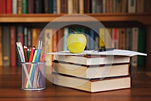 Stack of books on a desk with an apple and an organizer for pens