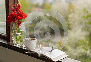 Book, glasses, cup of tea and red tulips on a wooden window. Read and rest.