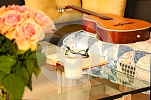 Book, glasses, roses and a mug with tea on modern glass table. An acoustic Spanish guitar is lying on an old rug on a black leathe