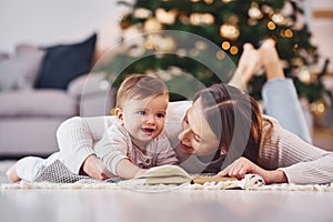 With book that is on the ground. Mother with her little daughter is indoors at home together